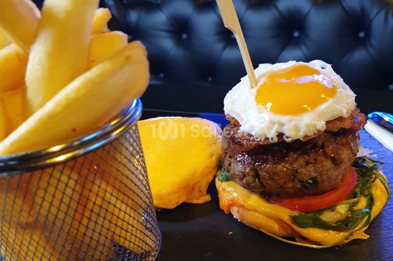 Burger garni d'un œuf au plat, bacon, légumes et fromage, accompagné de frites dans un panier métallique.