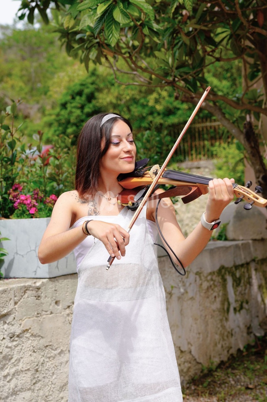 Une femme joue du violon en extérieur, entourée de verdure et de fleurs, près d'un muret en pierre.