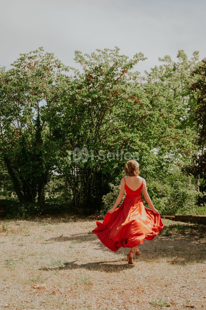 Une femme en robe rouge marche dans un jardin, entourée d'arbres et de verdure sous un ciel clair.
