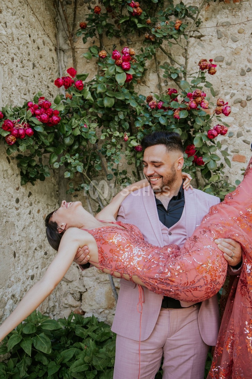 Un homme en costume rose porte une femme en robe scintillante devant un mur en pierre orné de rosiers fleuris.
