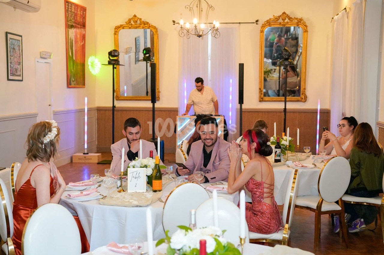 Des convives assis autour de tables décorées dans une salle élégante, avec un DJ en arrière-plan.