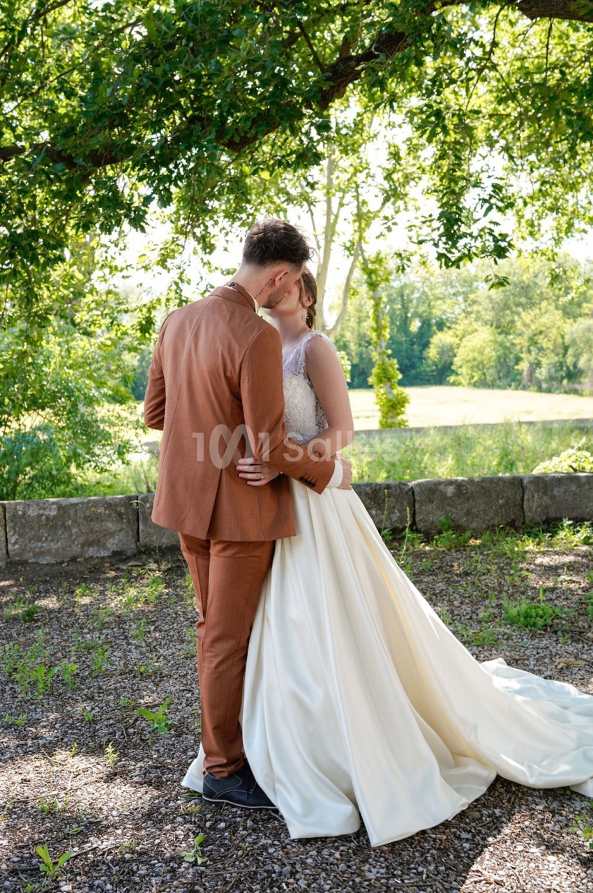 Un couple en tenue de mariage s'enlace sous un arbre, avec un paysage verdoyant en arrière-plan.