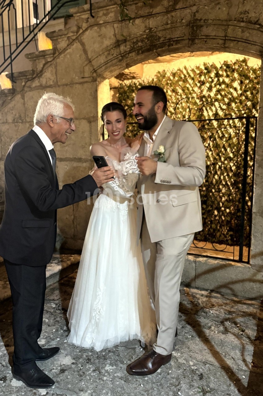 Un couple en tenue de mariage regarde un téléphone avec un homme âgé, debout devant un mur en pierre éclairé.