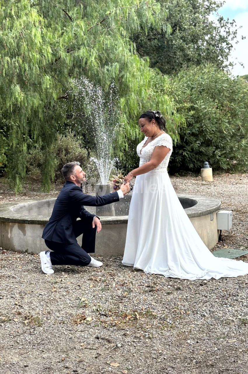 Un homme en costume propose à une femme en robe blanche devant une fontaine dans un jardin.