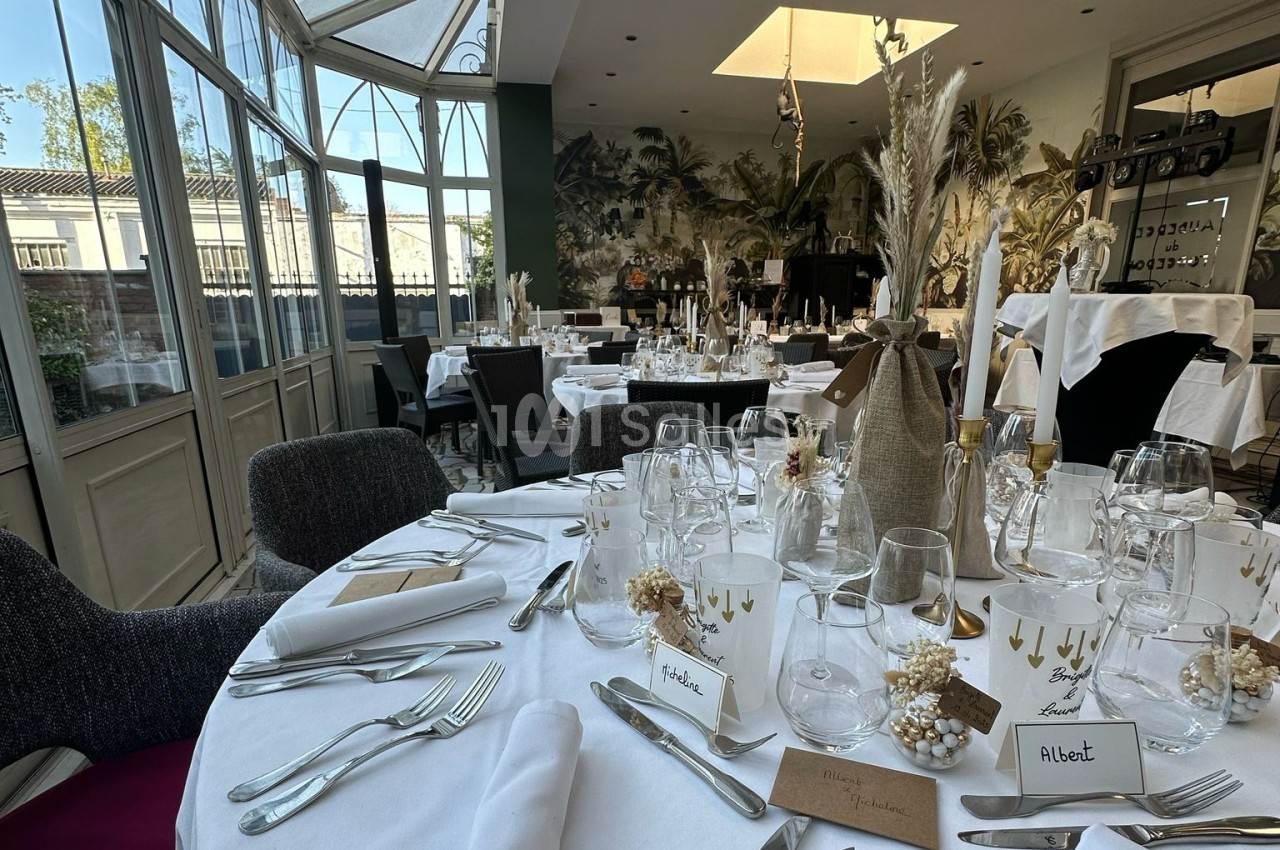 Salle de réception élégante avec tables rondes dressées, décorations florales et lumière naturelle provenant de grandes…
