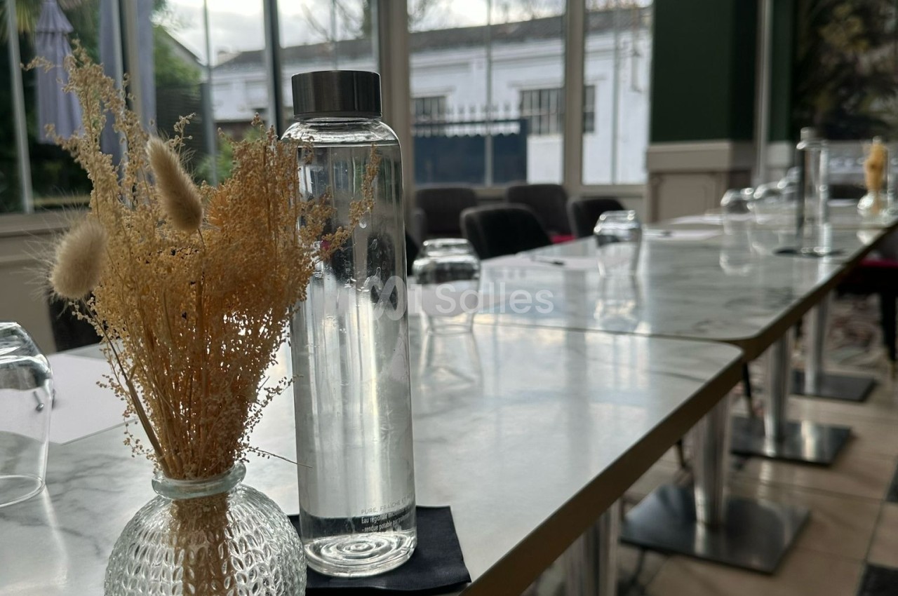 Bouteille d'eau et vase avec fleurs séchées sur une table en marbre dans une salle lumineuse avec grandes fenêtres.