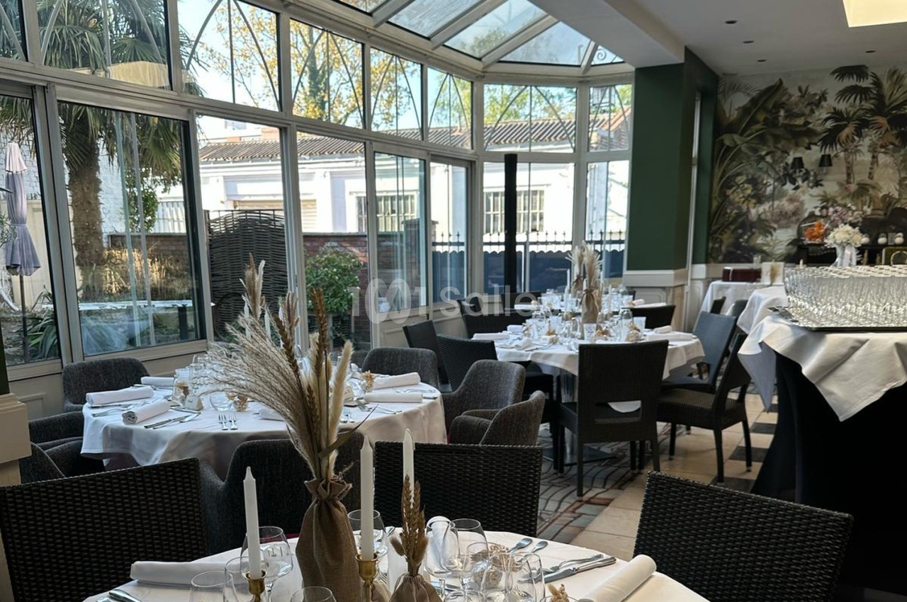 Salle lumineuse avec verrière, tables dressées avec nappes blanches, décorations florales et chaises en osier.