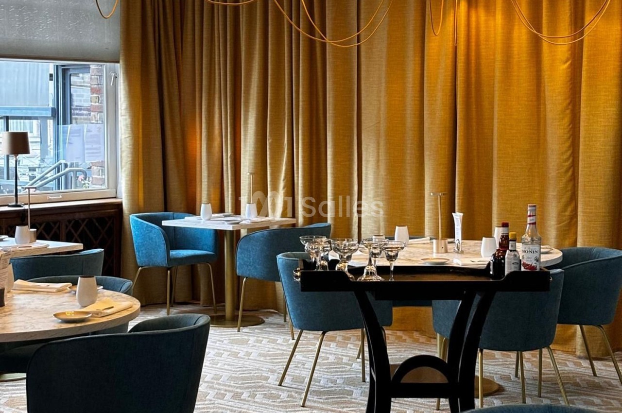 Salle de restaurant élégante avec tables dressées, chaises bleues, rideaux dorés et lumière tamisée.