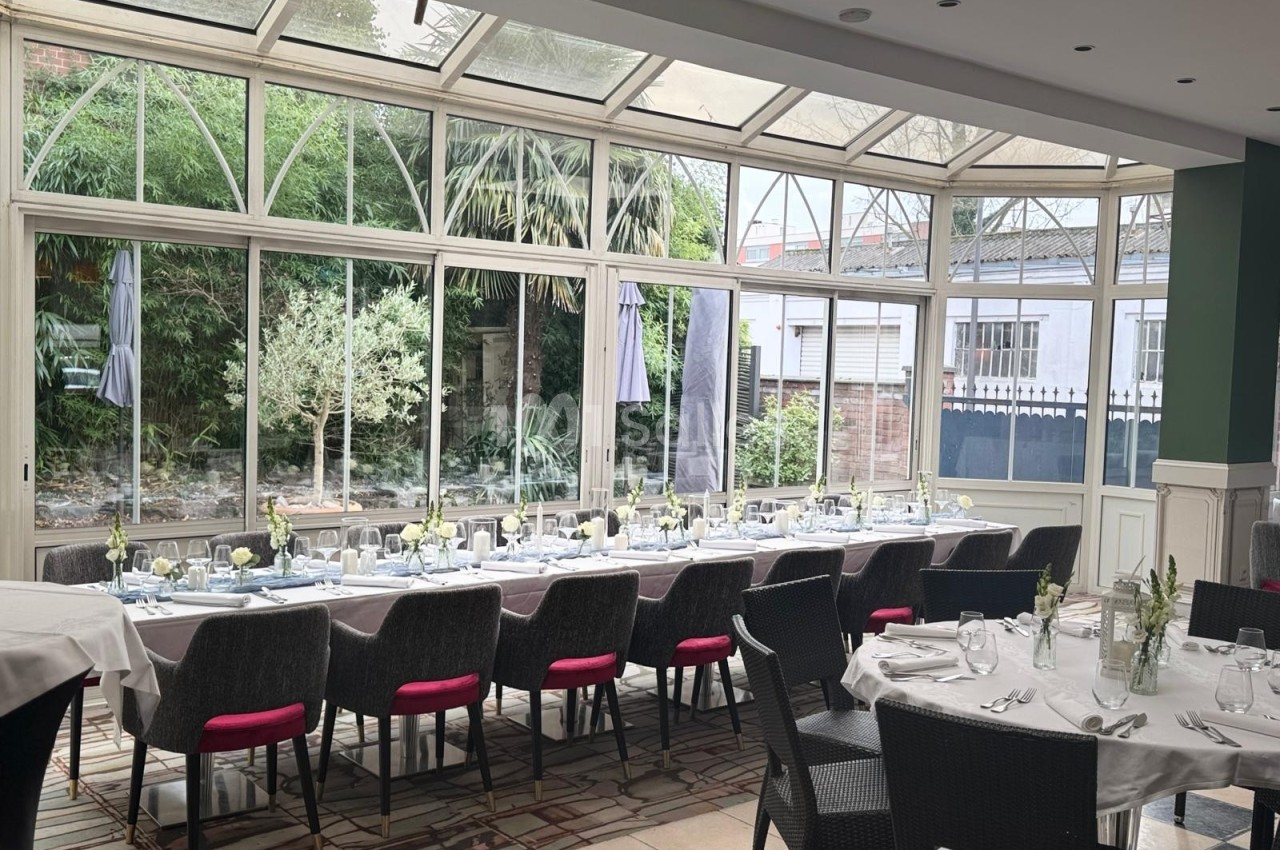 Salle lumineuse avec de grandes baies vitrées, tables dressées pour un repas, vue sur un jardin extérieur.