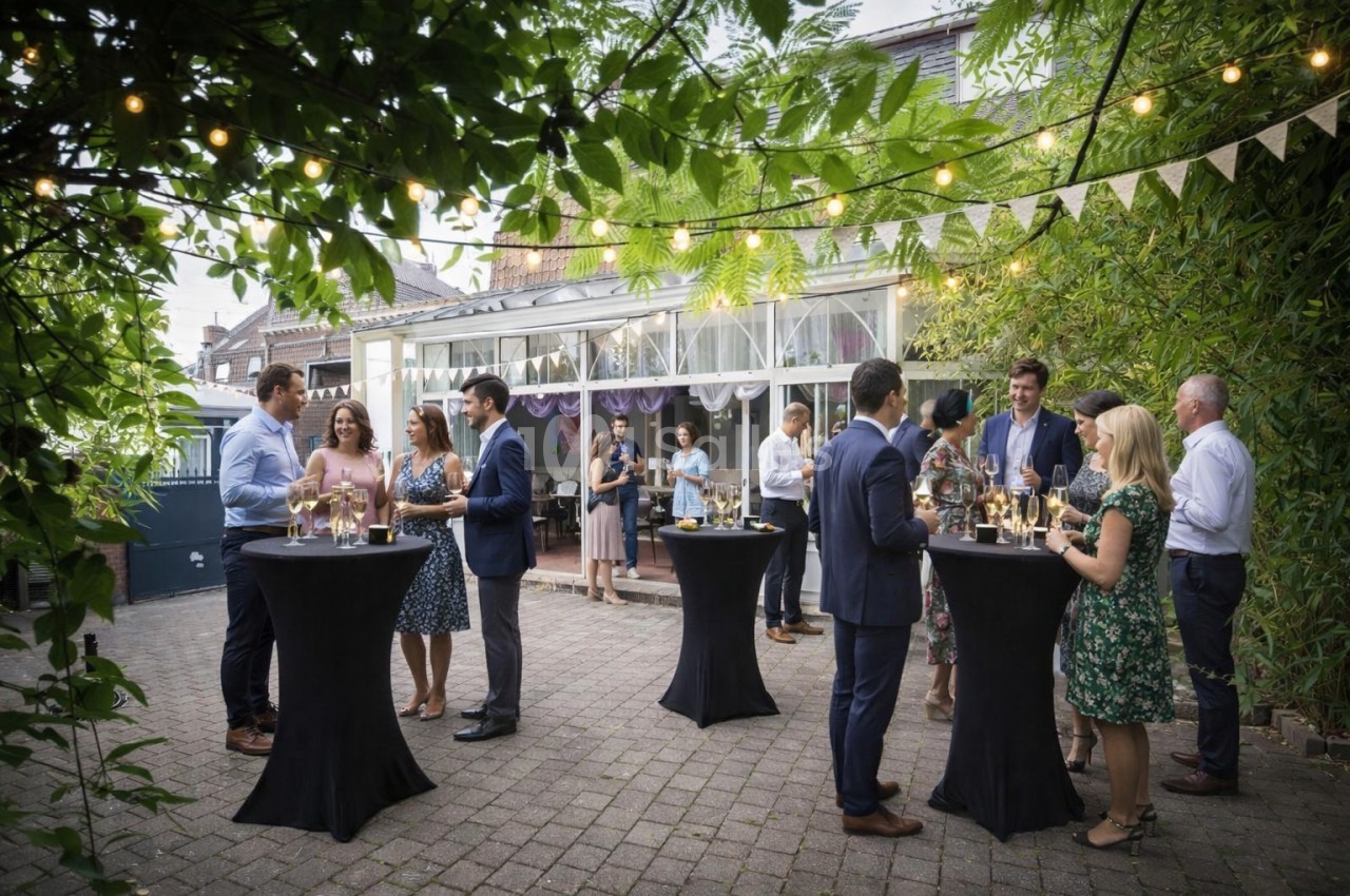Des personnes discutent en petits groupes autour de tables hautes lors d'un événement en extérieur sous des guirlandes…