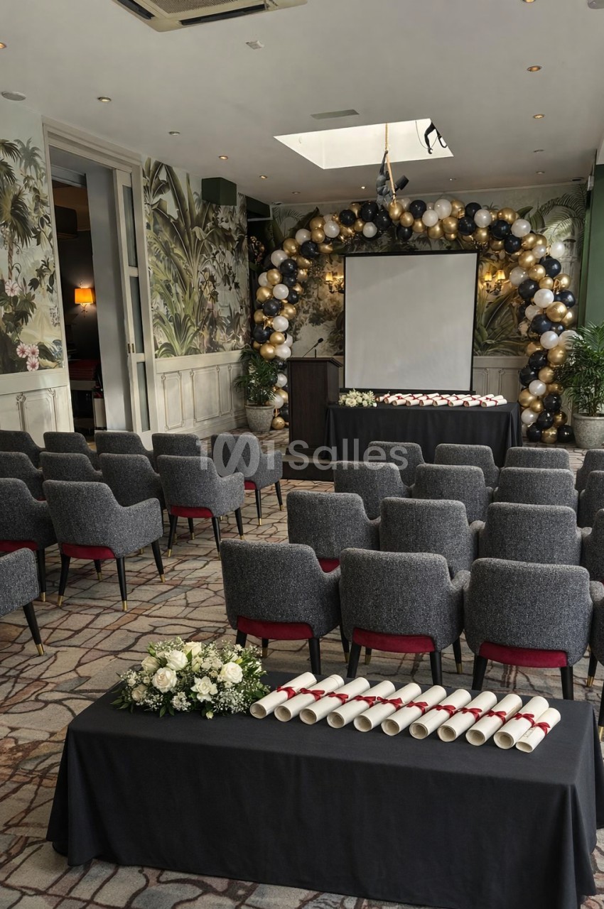 Salle décorée avec des ballons, écran de projection, chaises alignées et table avec diplômes et fleurs au premier plan.