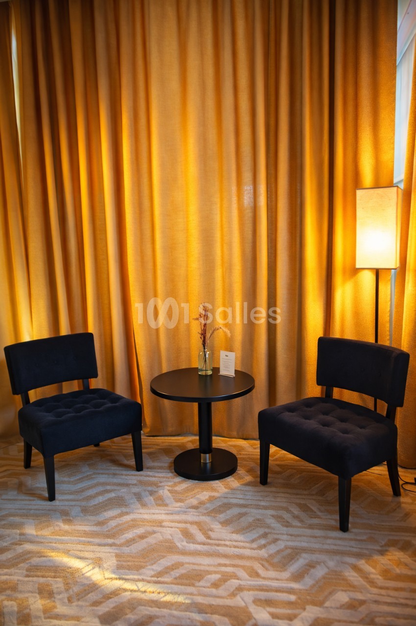 Deux fauteuils noirs entourent une table ronde avec un vase, devant des rideaux dorés et une lampe allumée.