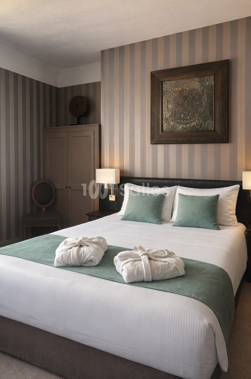 Chambre avec lit double, draps blancs, coussins verts, peignoirs pliés, mur à rayures et éclairage doux.