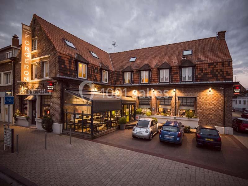 Façade d'un hôtel-restaurant en briques avec toit en tuiles, éclairé en soirée, et parking avec plusieurs voitures.