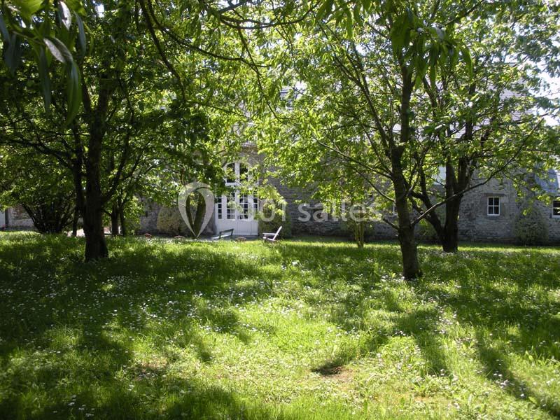 Jardin verdoyant avec des arbres ombrageant une pelouse, devant une maison en pierre aux fenêtres blanches.