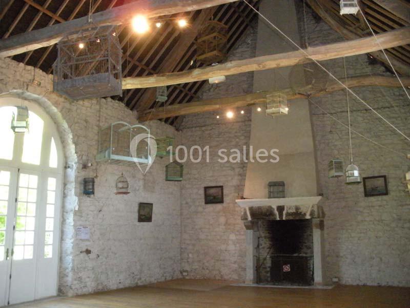Salle en pierre avec cheminée, poutres apparentes et cages à oiseaux suspendues au plafond.