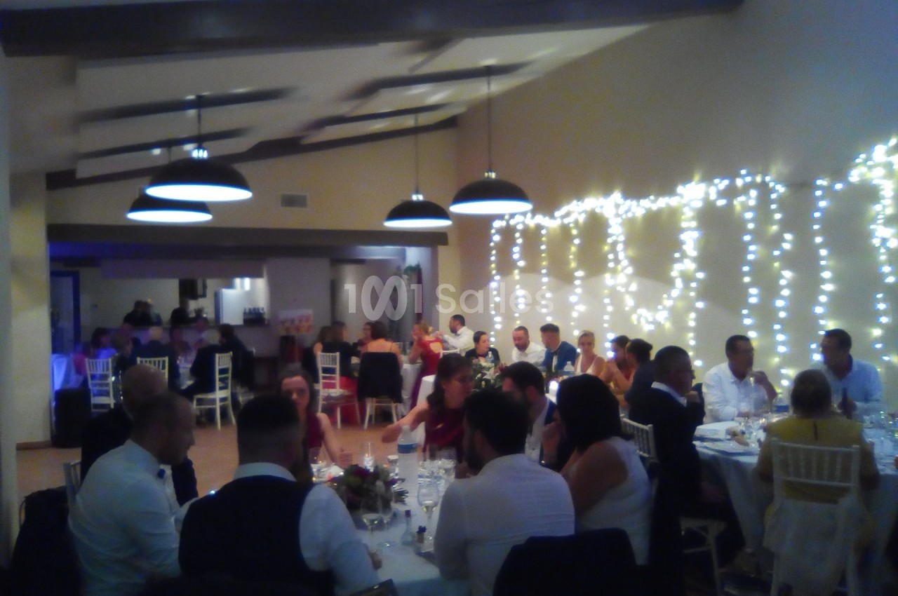 Salle de réception avec des invités assis à des tables, éclairée par des guirlandes lumineuses et des suspensions noires.