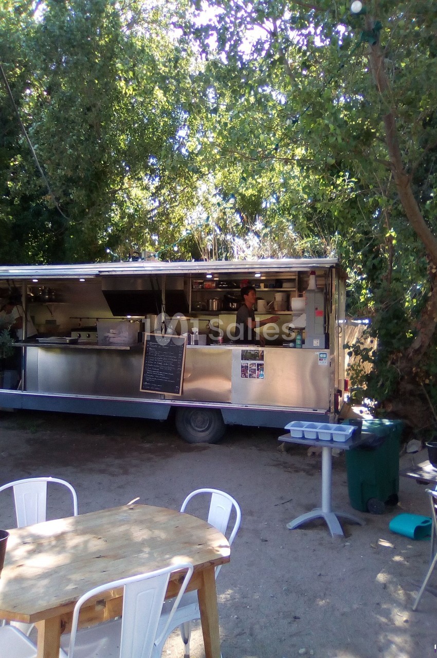 Camion de restauration mobile en plein air, entouré d'arbres, avec une table et des chaises au premier plan.