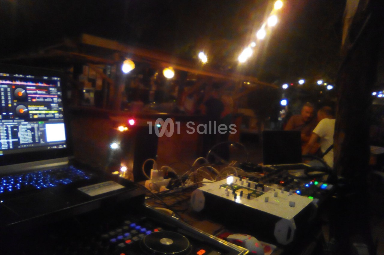 Poste de DJ avec ordinateurs et tables de mixage, éclairé par des guirlandes lumineuses dans un espace extérieur nocturne.