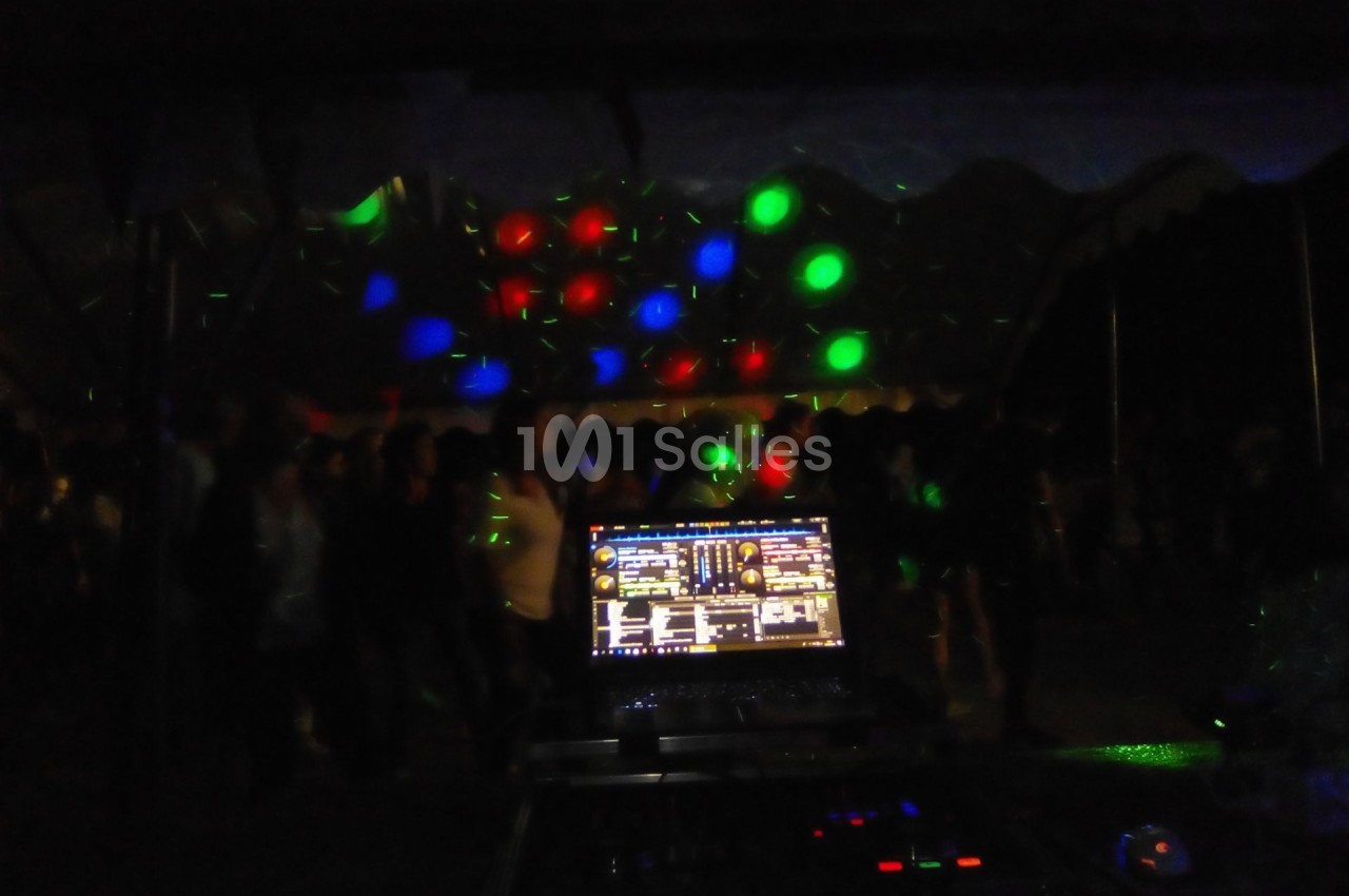 Vue d'une console de DJ éclairée, avec des lumières colorées projetées sur une foule dans un espace sombre.