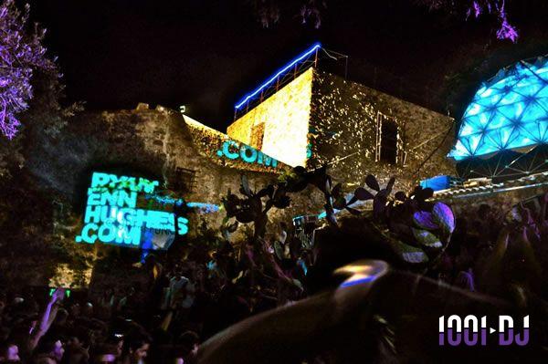 Foule dansante en soirée extérieure avec projections lumineuses sur des murs en pierre et une structure géométrique éclairée.