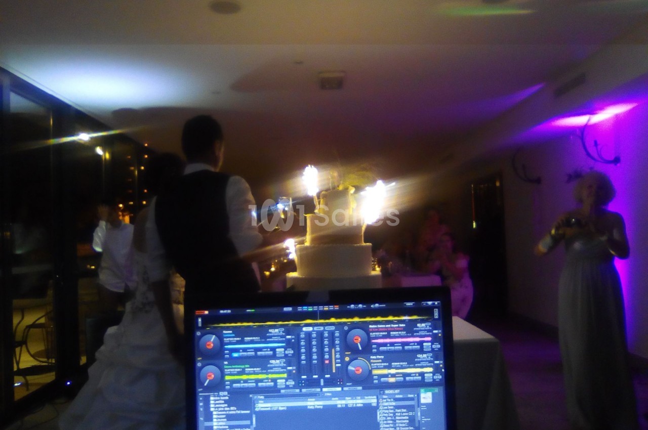 Un DJ mixe lors d'une réception de mariage, avec une table de mixage en premier plan et un gâteau illuminé en arrière-plan.