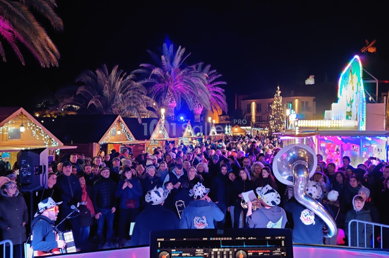 Foule rassemblée devant des stands illuminés lors d'un événement nocturne, avec un DJ en premier plan.