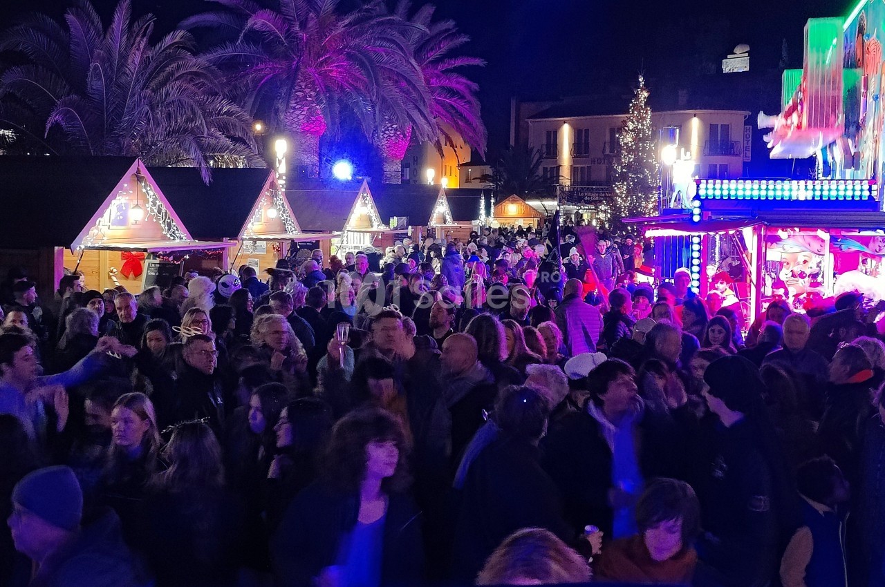Foule rassemblée de nuit dans un marché de Noël illuminé, avec chalets décorés et ambiance festive.