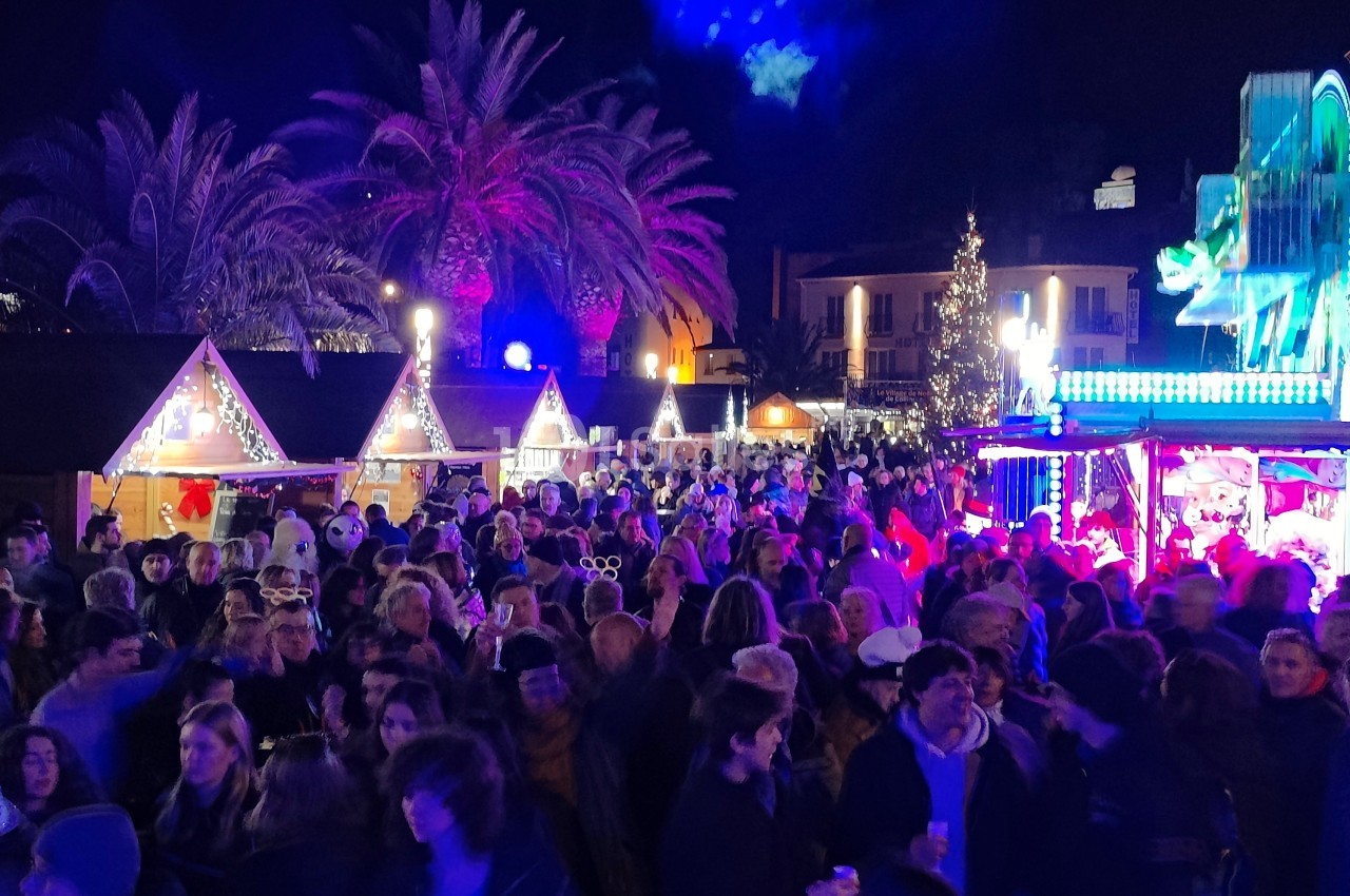 Foule rassemblée dans un marché de Noël nocturne éclairé par des lumières colorées, avec des stands et un sapin décoré.
