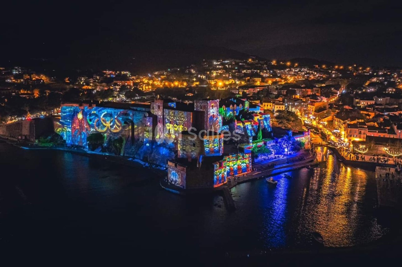 Vue nocturne d'une ville côtière illuminée avec des projections colorées sur un château et des lumières dans les rues.