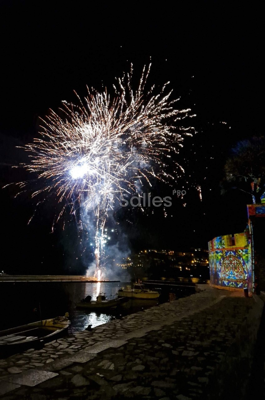 Feu d'artifice illuminant le ciel nocturne au-dessus d'un quai avec des barques et une façade colorée éclairée.