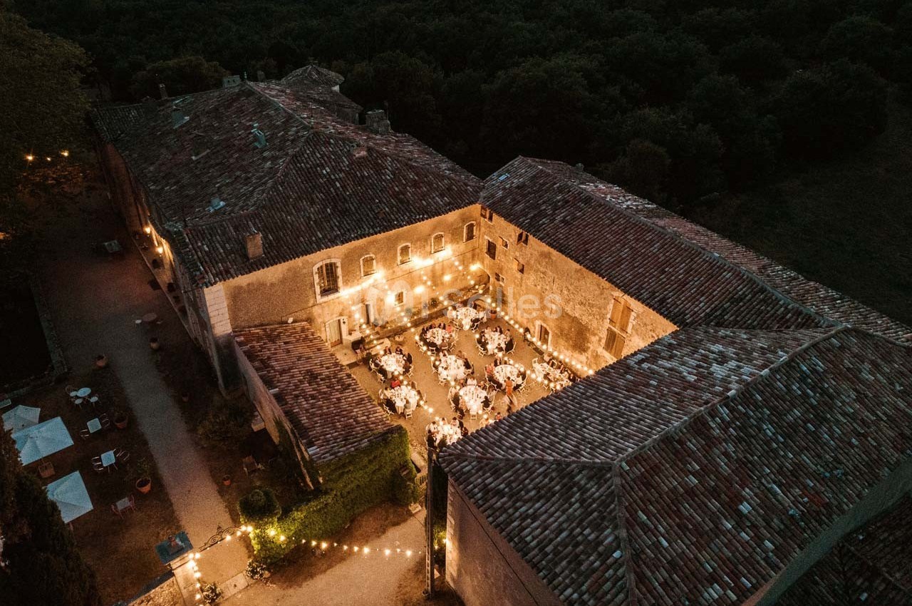 Vue aérienne d'une cour intérieure éclairée pour un dîner en plein air, entourée de bâtiments en pierre et de végétation.