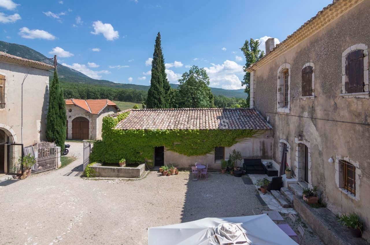 Cour d'une maison en pierre avec murs recouverts de lierre, entourée de bâtiments anciens et vue sur des collines…