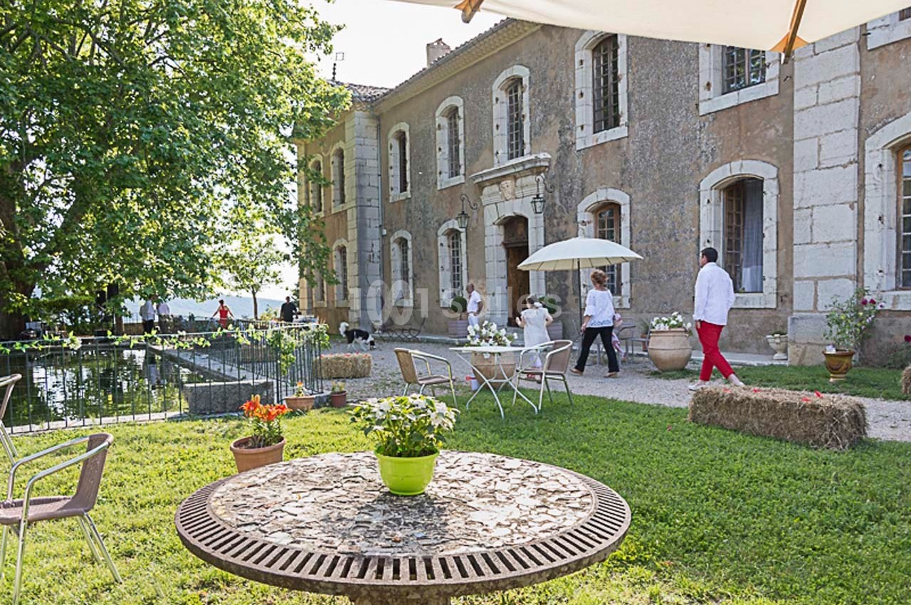 Terrasse d'un domaine ancien avec des tables, des chaises et des visiteurs près d'un bassin entouré de verdure.