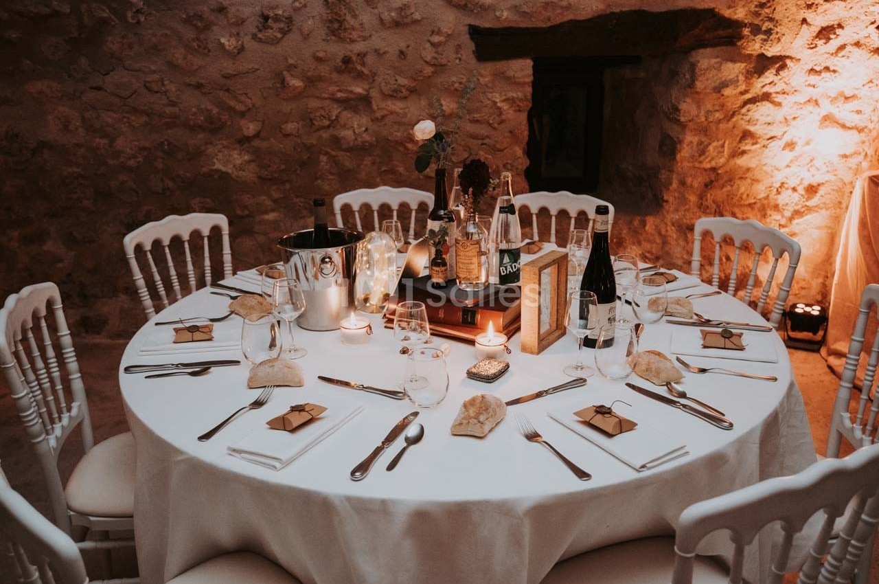 Table ronde décorée pour un dîner, avec nappes blanches, couverts, bougies et cadeaux sur chaque assiette.