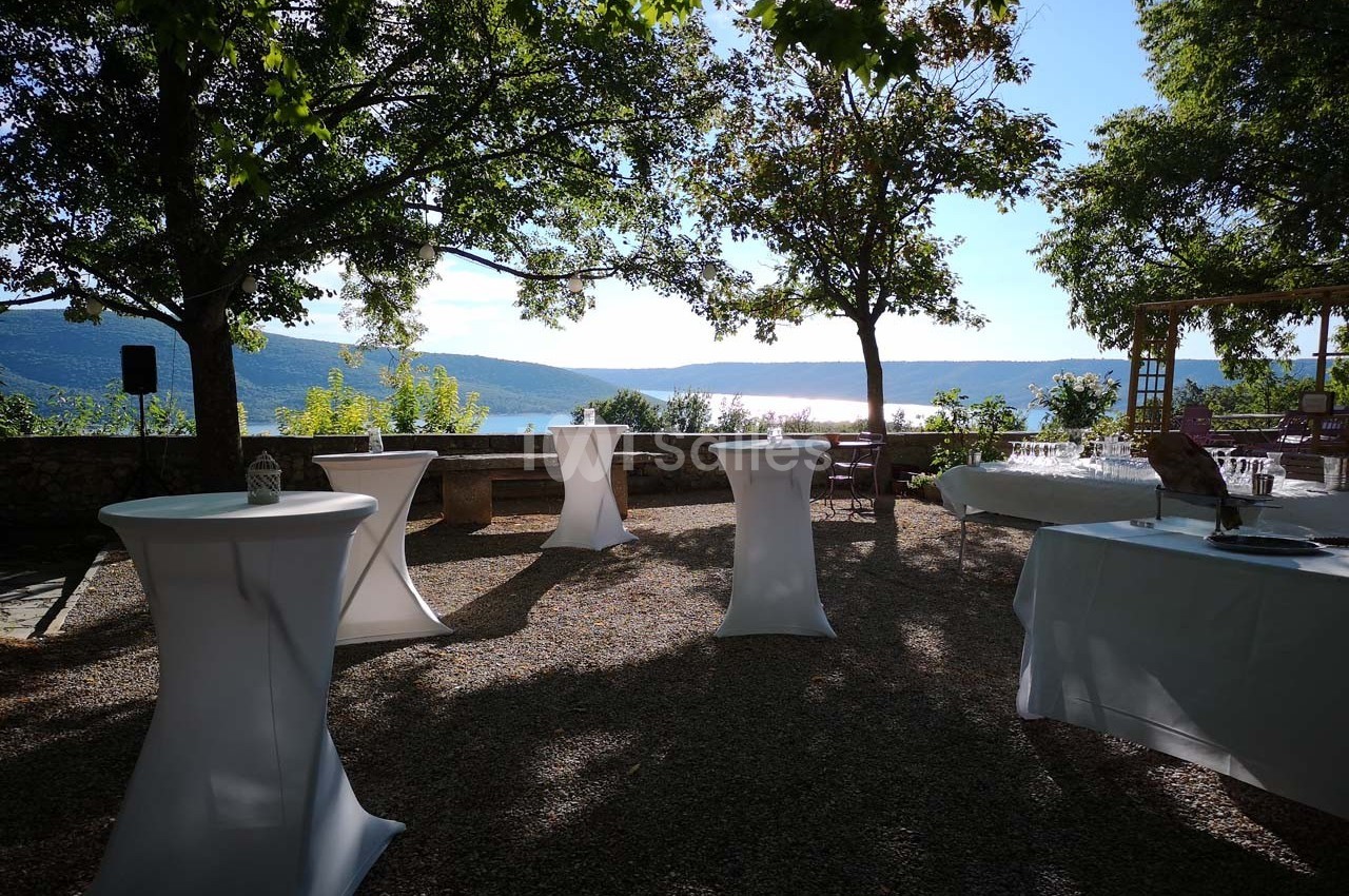 Tables hautes dressées en extérieur sous des arbres, avec vue sur un lac et des collines en arrière-plan.