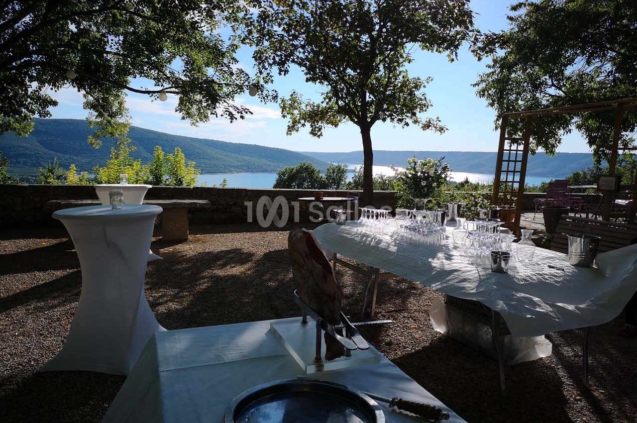 Vue d'une terrasse ombragée avec tables dressées, offrant un panorama sur un lac entouré de collines verdoyantes.