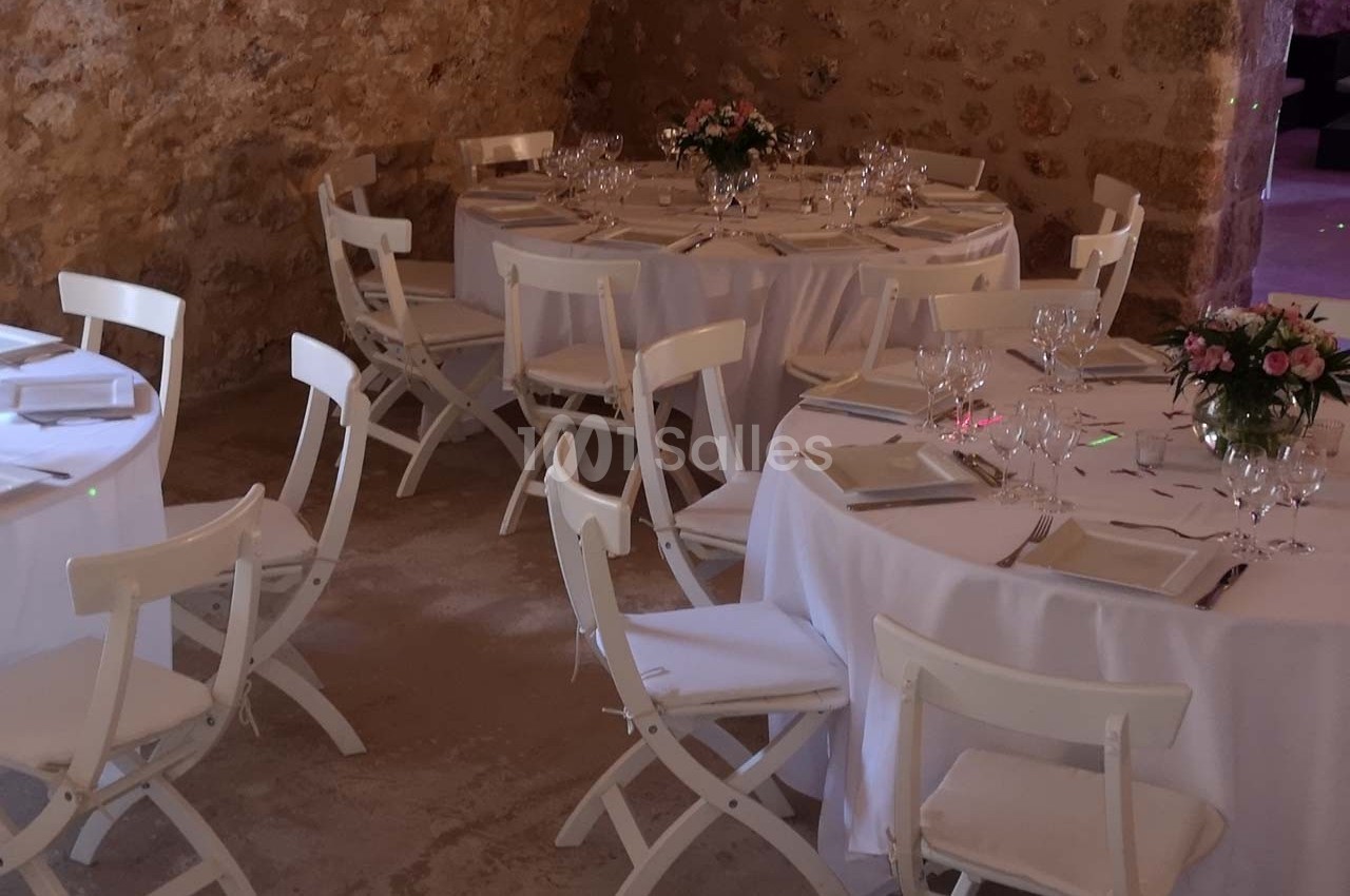 Salle en pierre aménagée avec des tables rondes dressées de nappes blanches, vaisselle et bouquets de fleurs.