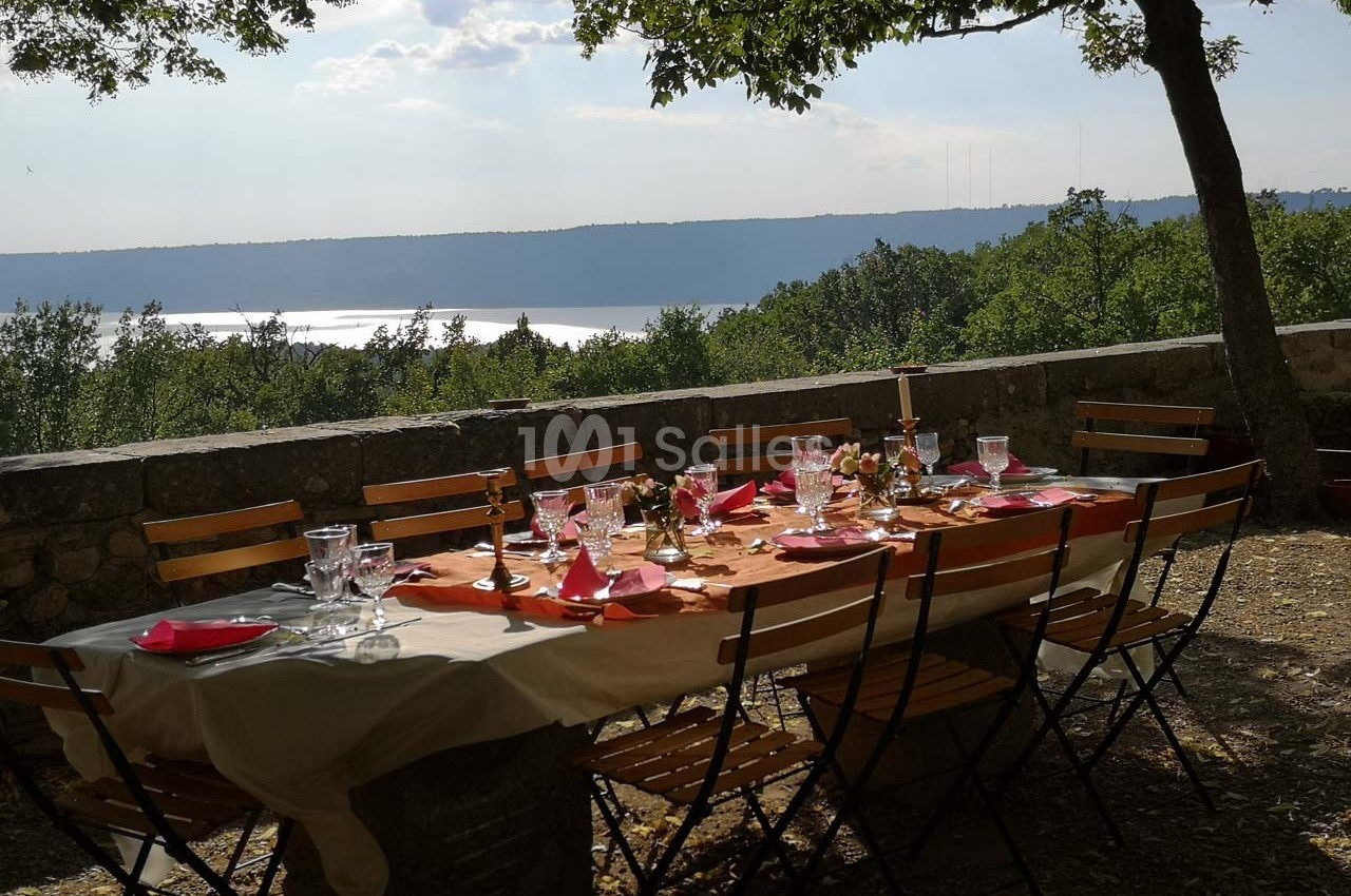 Table dressée en extérieur avec nappes, vaisselle et décorations, sous des arbres, vue sur un lac et des collines.