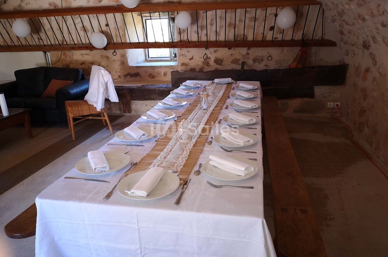 Table en bois dressée avec une nappe blanche, des assiettes, des serviettes pliées et des couverts, dans une salle rustique.
