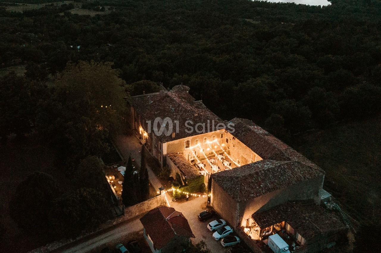 Vue aérienne d'un domaine en pierre éclairé la nuit, entouré de végétation et de voitures stationnées.