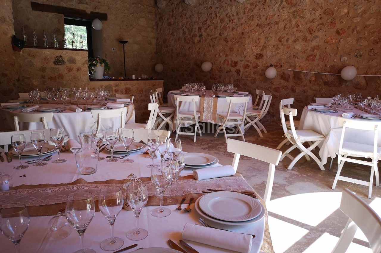 Salle en pierre avec tables rondes et rectangulaires dressées pour un repas, éclairée par la lumière naturelle.