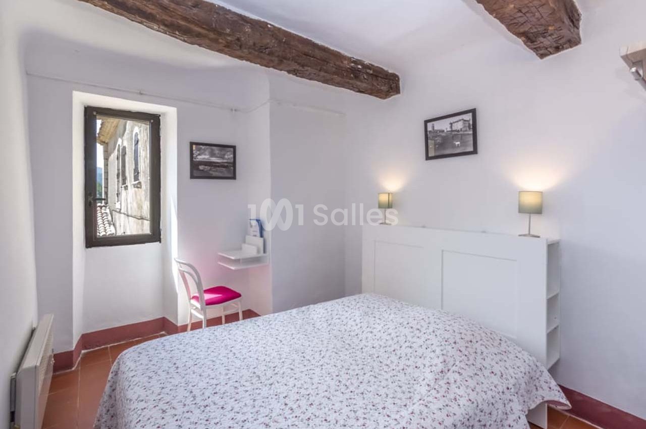Chambre lumineuse avec lit double, poutres apparentes, bureau près d'une fenêtre et deux lampes de chevet allumées.