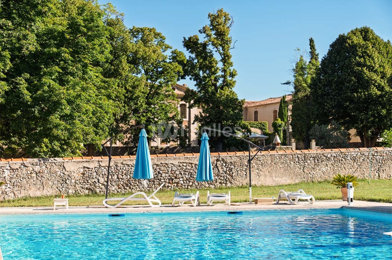 Piscine extérieure entourée de chaises longues, parasols bleus et arbres, avec un bâtiment en arrière-plan.