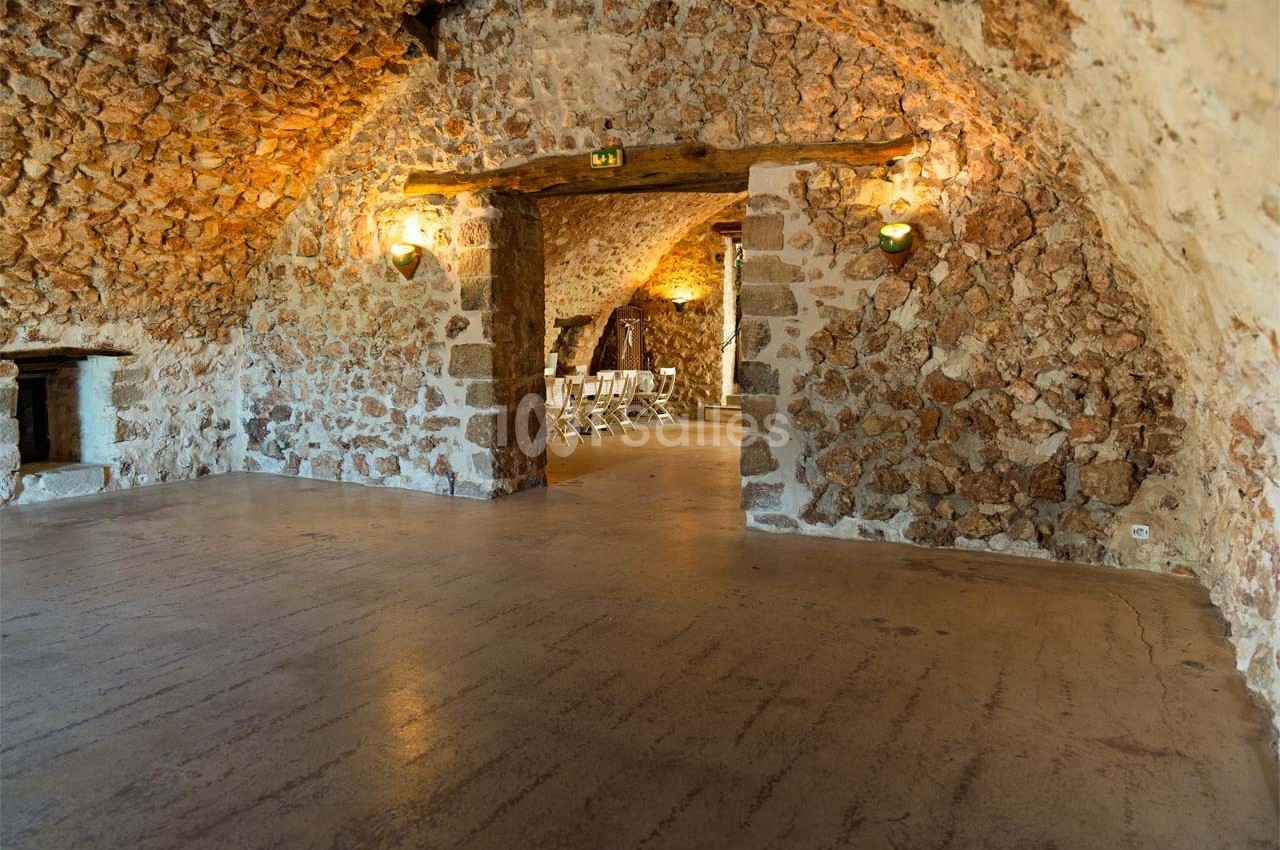 Salle voûtée en pierre avec éclairage mural, sol en béton et ouverture donnant sur des chaises alignées.