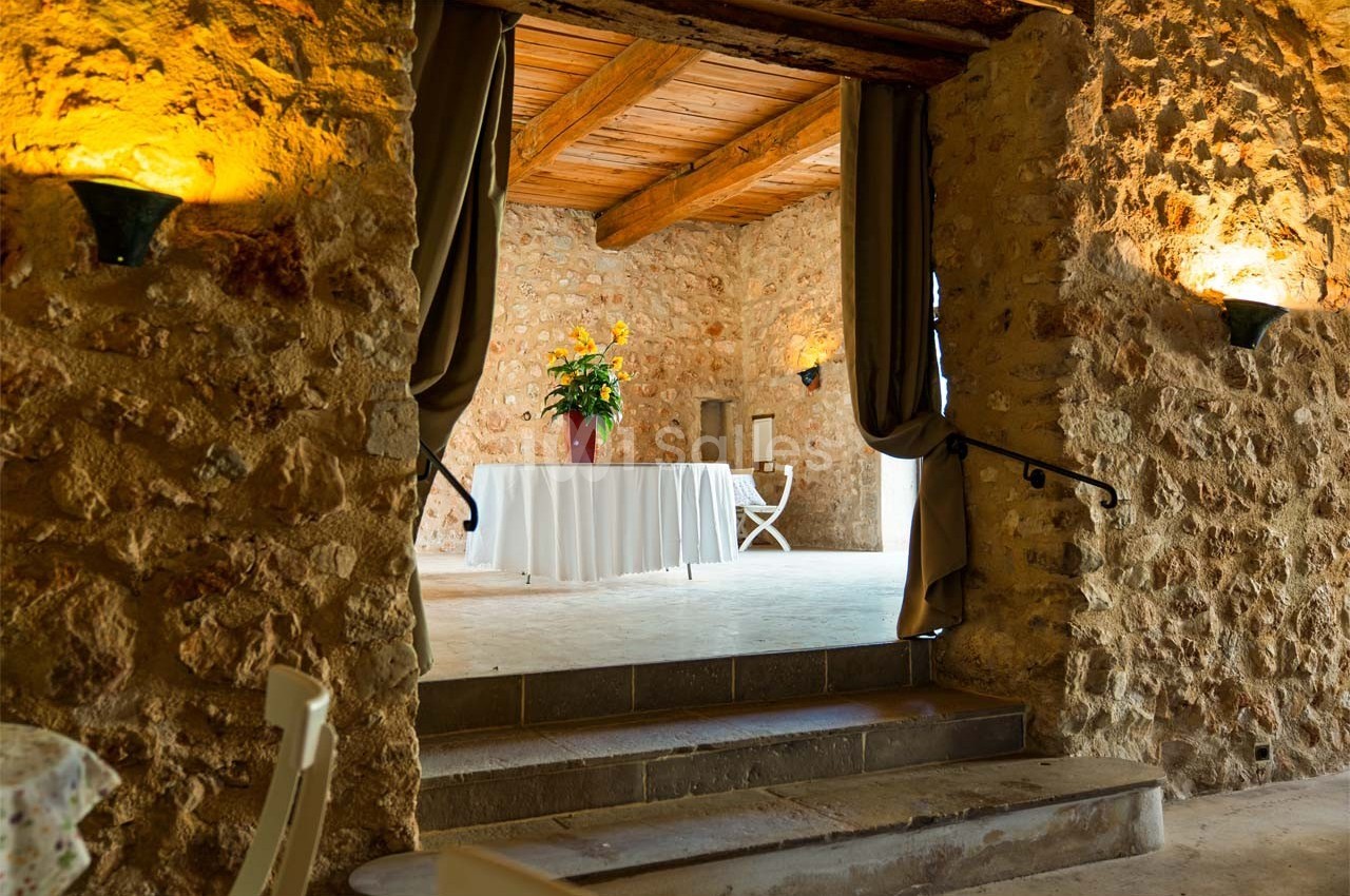Salle en pierre avec plafond en bois, table blanche décorée de fleurs jaunes et rideaux encadrant l'entrée.