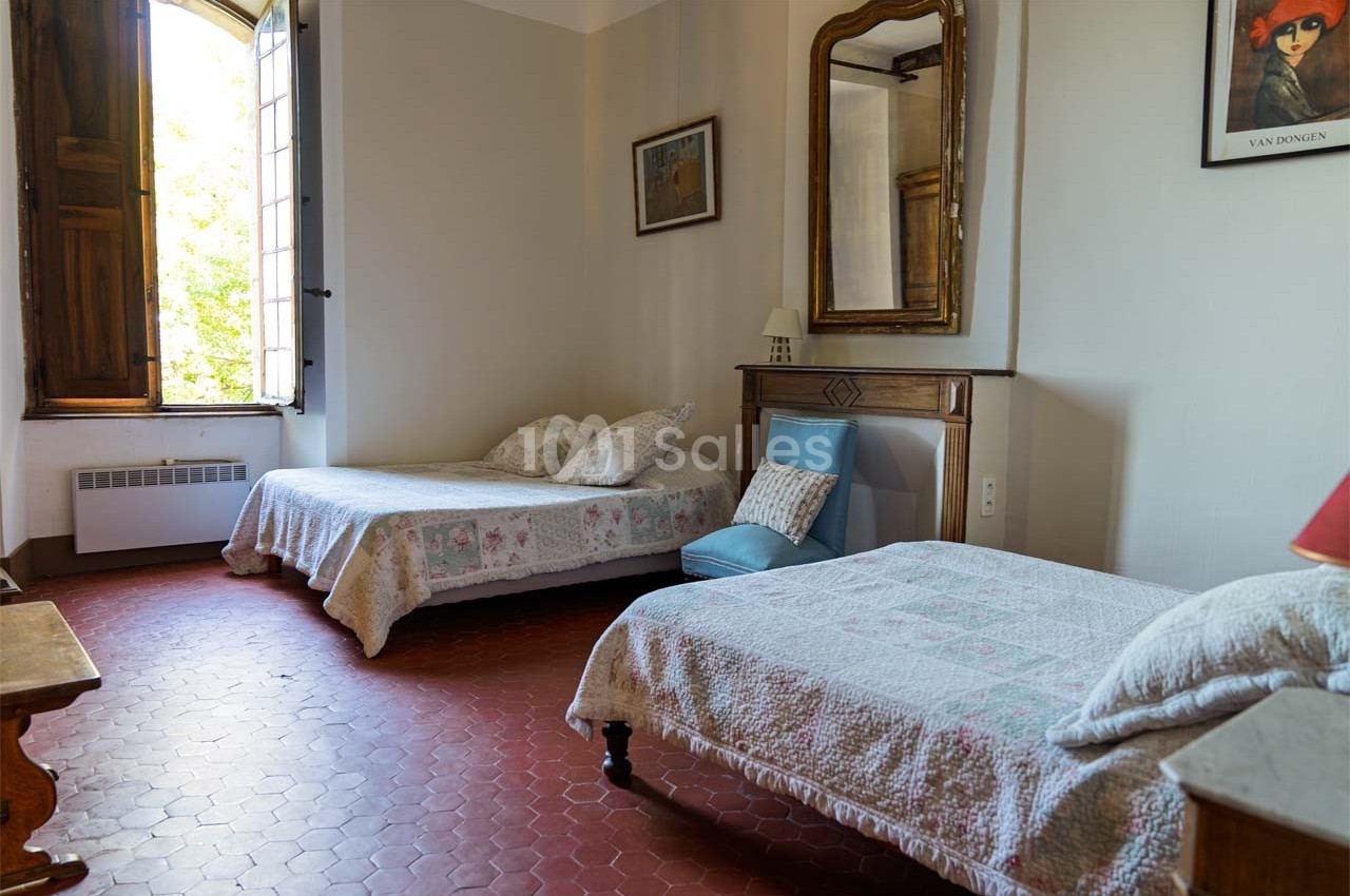 Deux lits simples avec couvre-lits blancs dans une chambre lumineuse au sol en terre cuite, avec une fenêtre ouverte.