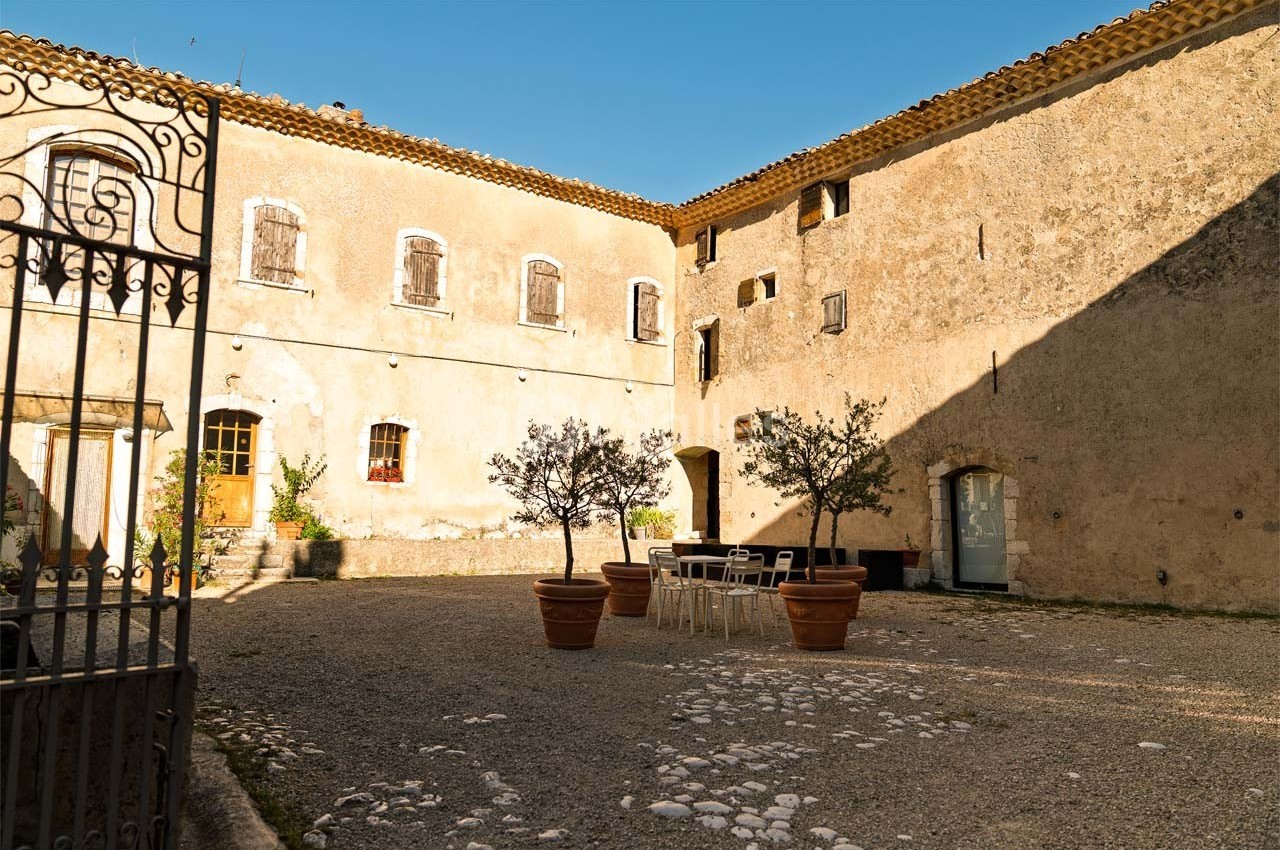 Cour intérieure ensoleillée avec des murs en pierre, des pots d'oliviers et une table entourée de chaises blanches.