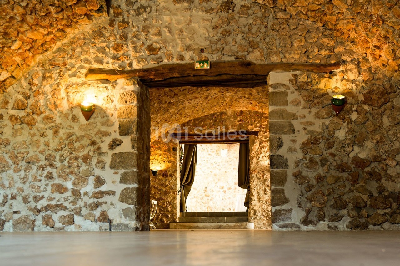 Salle voûtée en pierre avec éclairage mural, ouvrant sur une porte encadrée de rideaux et une lumière naturelle.