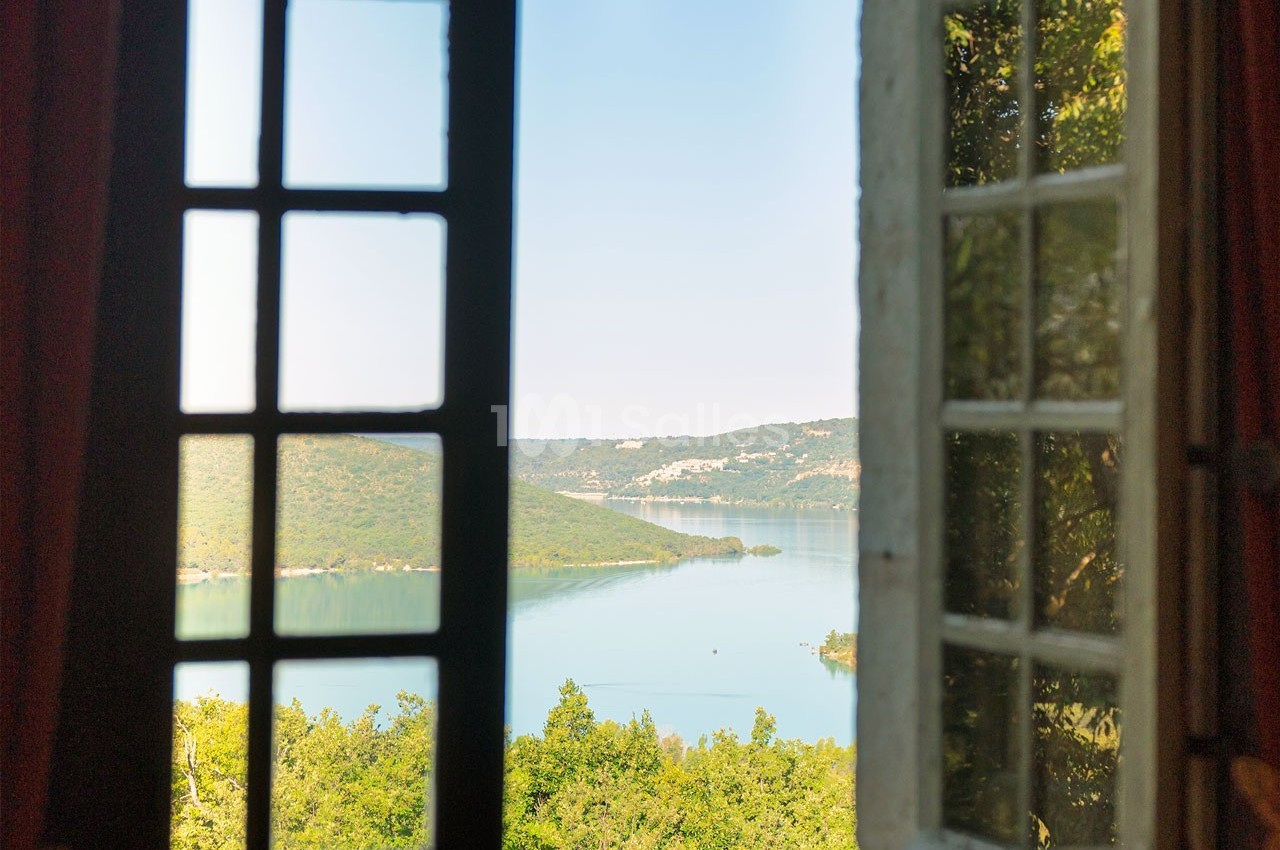 Vue sur un lac entouré de collines verdoyantes à travers une fenêtre ouverte avec des volets en bois.